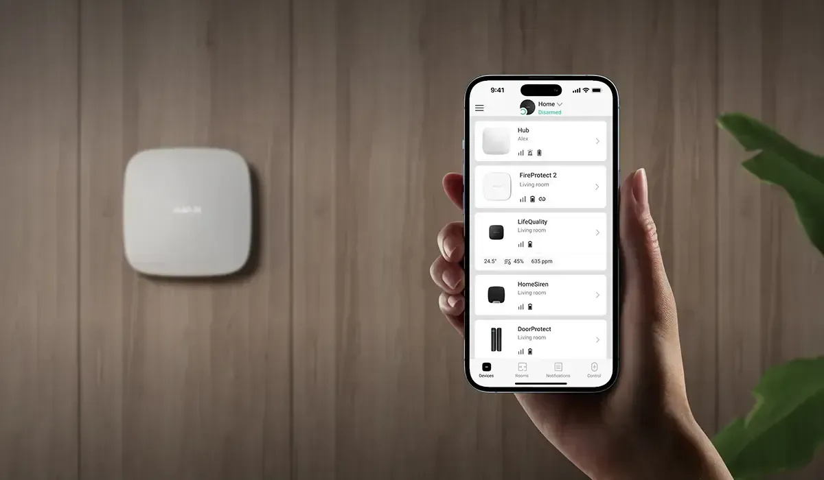 Hand holding smartphone displaying smart home device list next to mounted white access point.