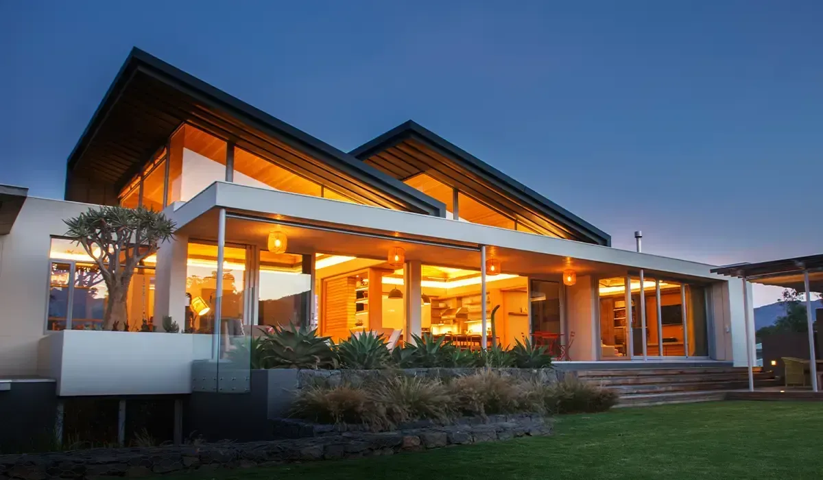 Modern house with large windows, glowing interior lights, and a grassy yard at dusk.