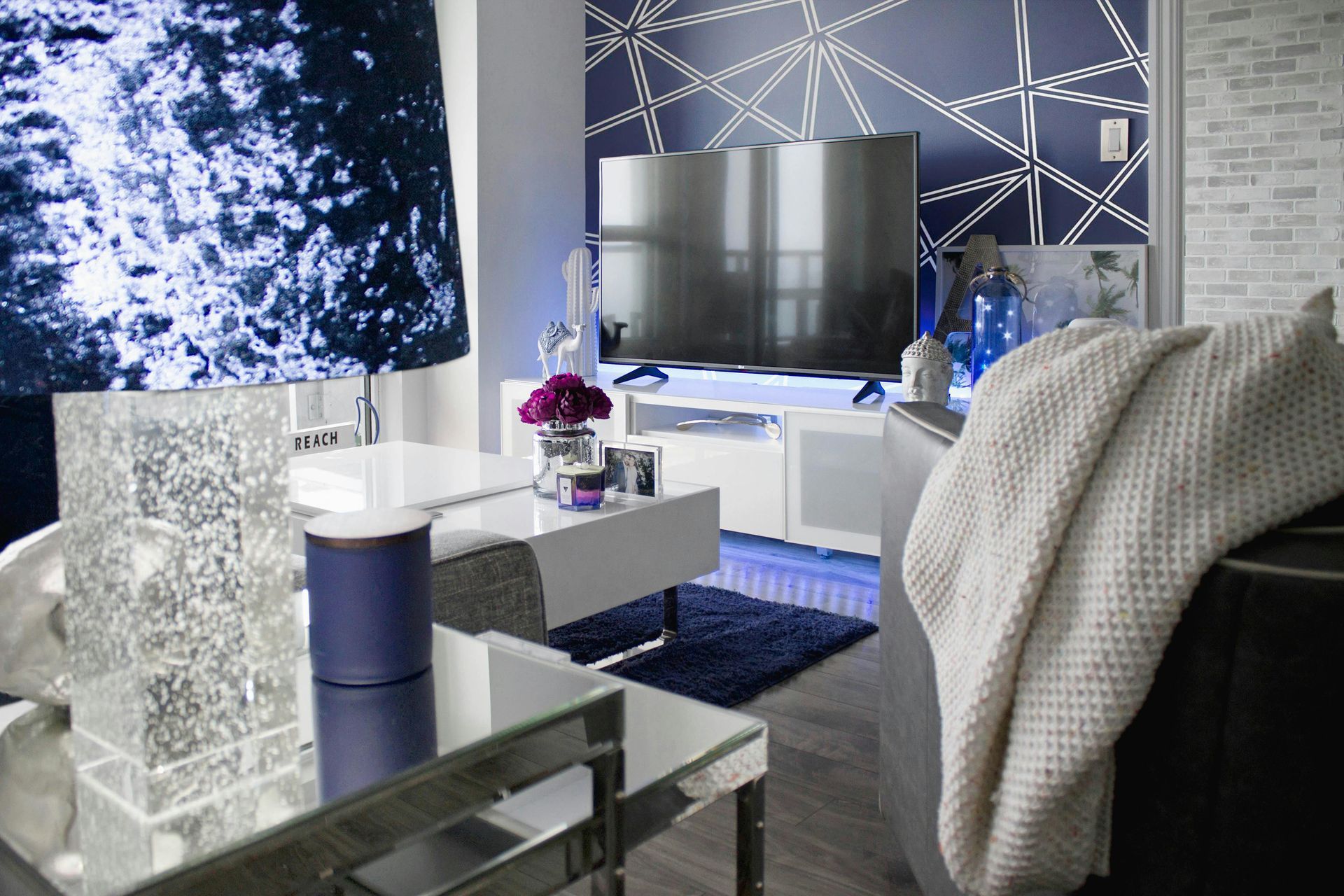 Modern living room with a large flat-screen TV, geometric accent wall, blue decorative lighting, and