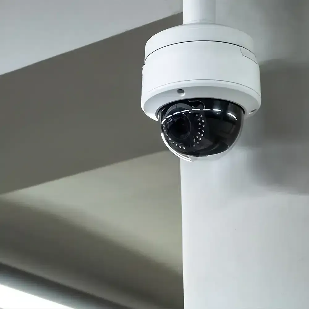 White security camera mounted on a white column, angled against a neutral gray wall.
