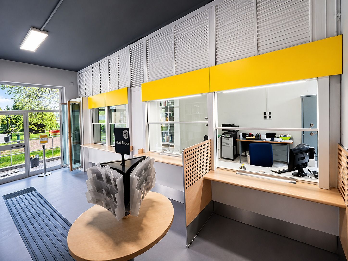 Una luminosa hall per uffici con un bancone di servizio rifinito in giallo, un tavolo espositivo in legno e un'area riservata al personale con pareti in vetro.