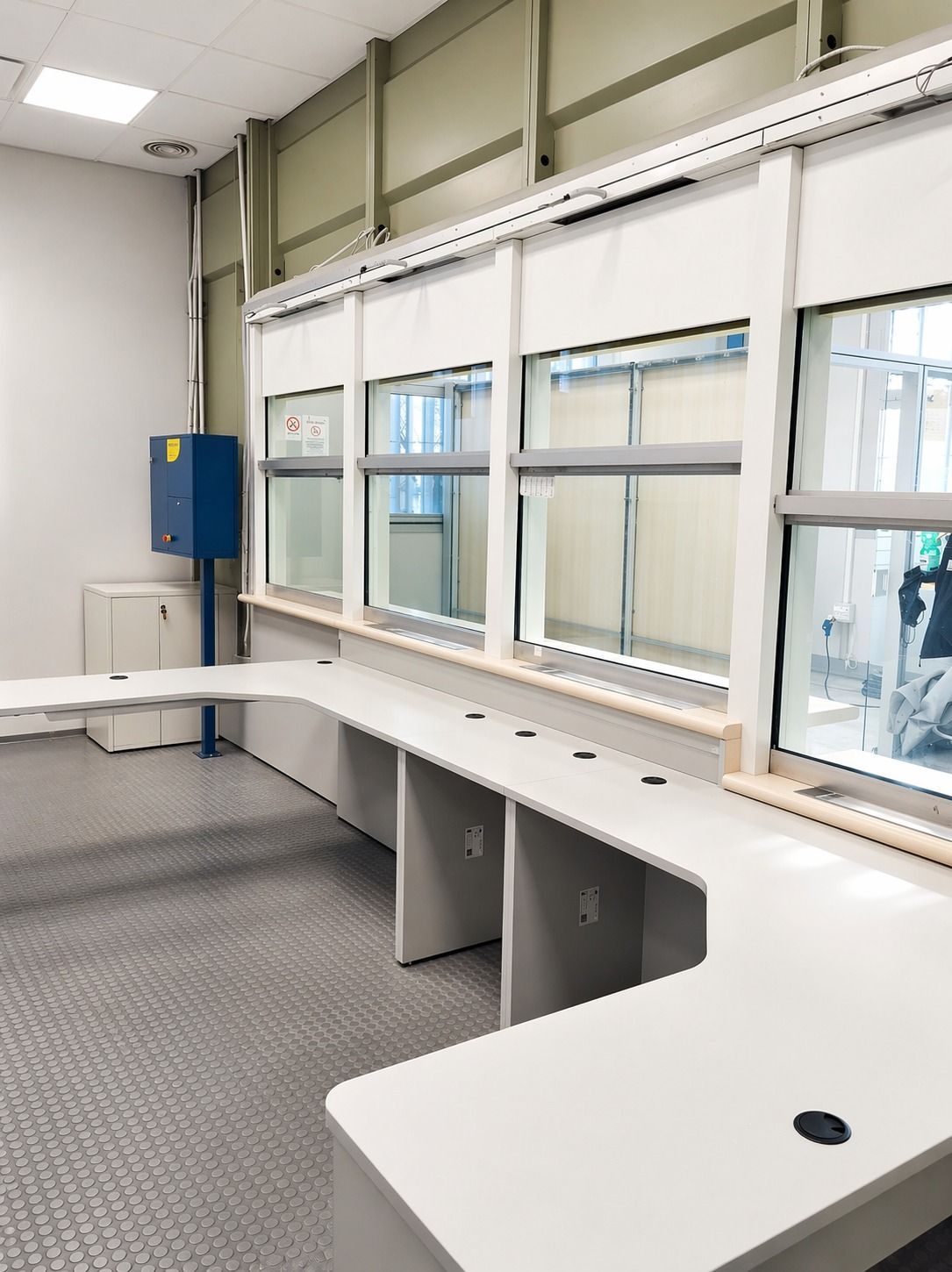 In una stanza luminosa e moderna, accanto a una parete con ampie vetrate e a un mobile blu, si trova una scrivania bianca a forma di L con passacavi.