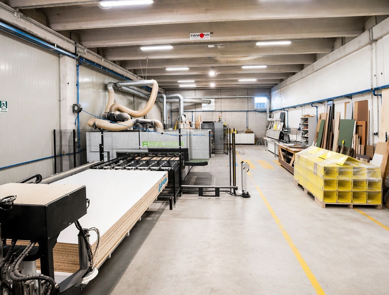 Un'officina di falegnameria luminosa e pulita, con un grande tavolo da lavoro bianco, macchinari pesanti e contenitori gialli per i materiali.