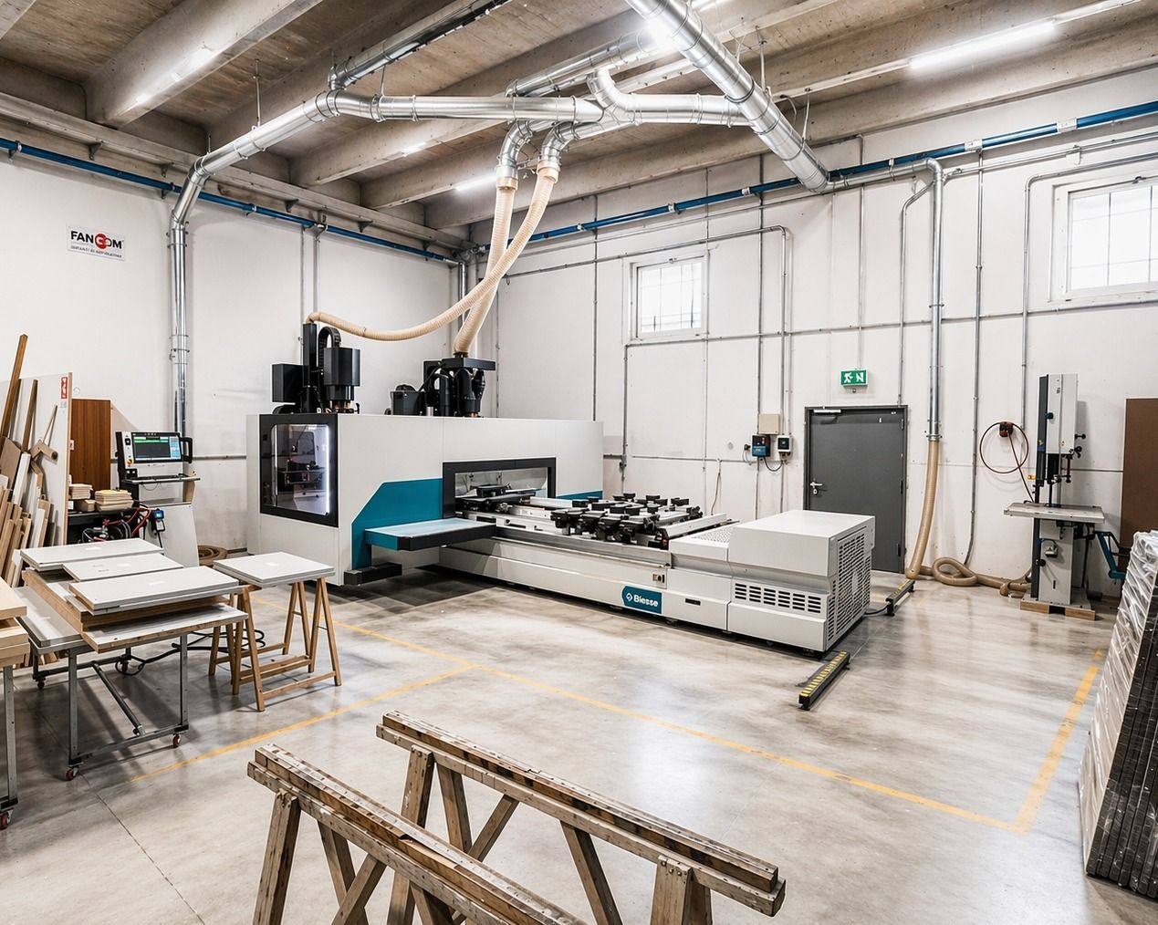 In un'officina industriale pulita, con tubi di aspirazione della polvere e cavalletti di legno nelle vicinanze, si trova una grande macchina per la lavorazione del legno a controllo numerico.