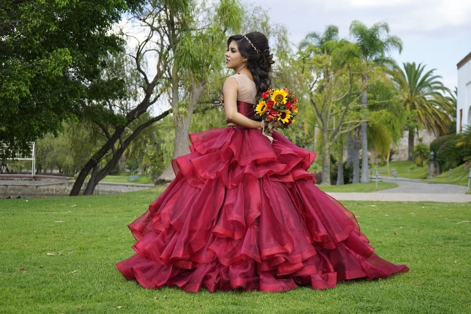 A person in a voluminous dark red ballgown holds a bouquet of sunflowers while standing in a grassy park setting.