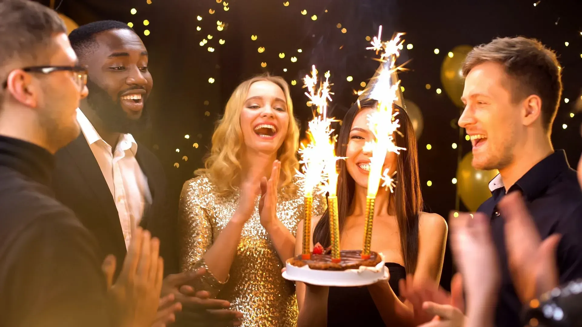 Friends cheer and clap as a woman holds a birthday cake with lit sparklers in a room decorated with gold lights.