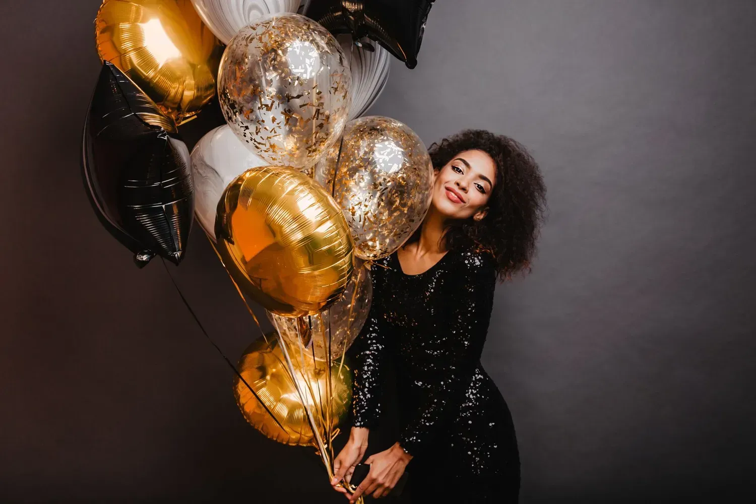 A smiling person in a sequined black dress poses against a dark backdrop while holding a large bunch of gold balloons.