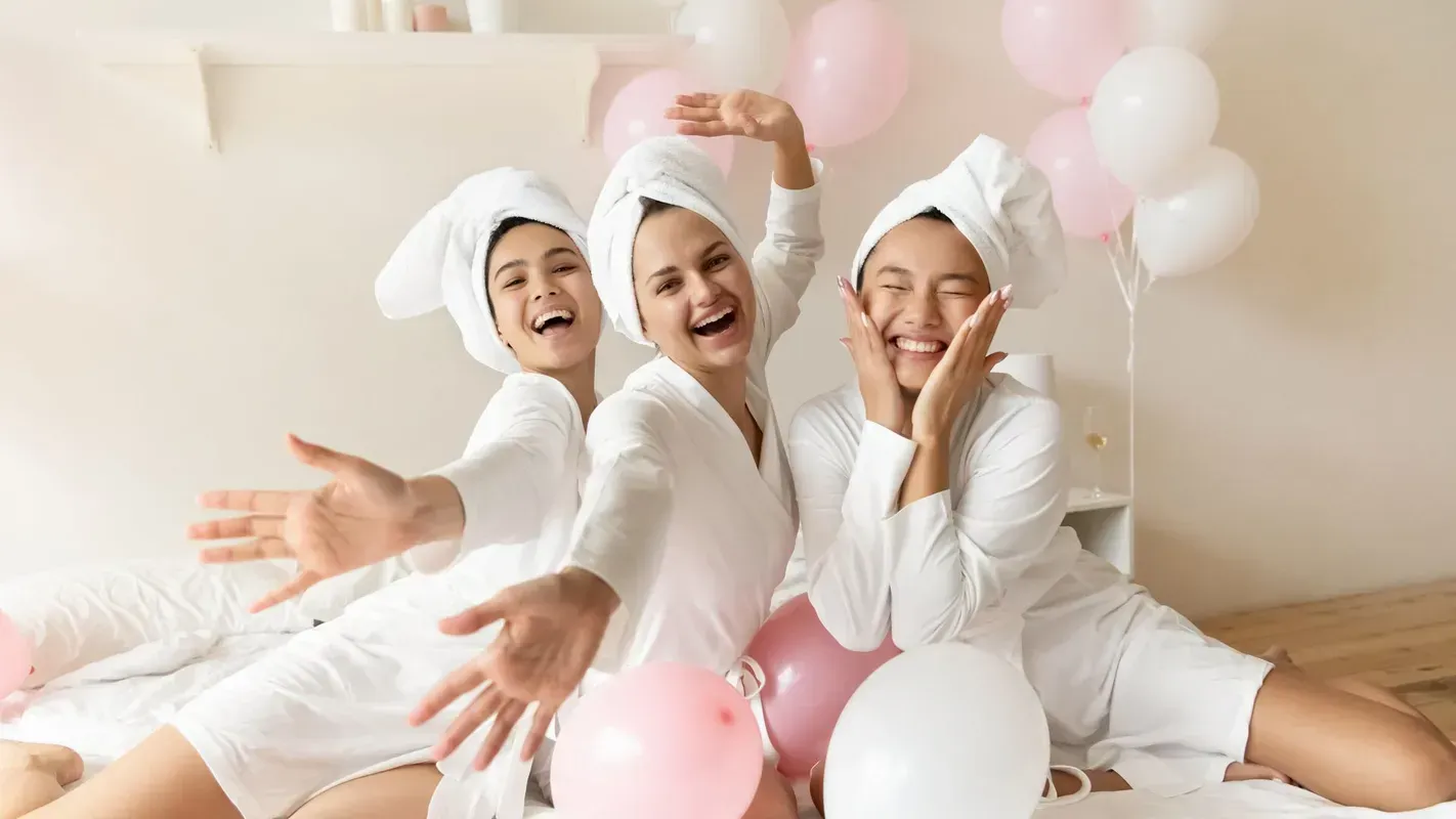 Three people in white robes and head towels pose playfully on a bed with pink and white balloons in a bright room.