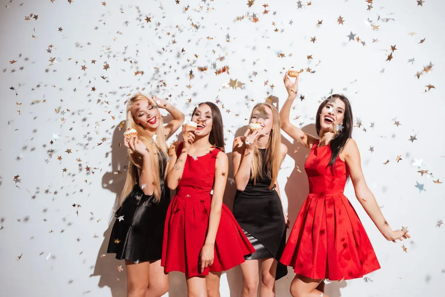 Four women in red and black dresses laugh and eat cake while golden confetti falls against a white wall.