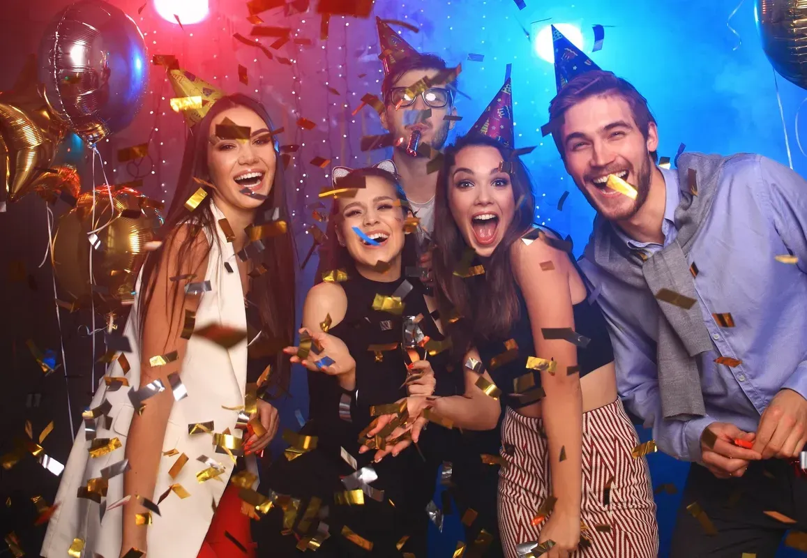 Four friends wearing party hats cheer while surrounded by falling gold confetti in a room lit with blue and red lights.