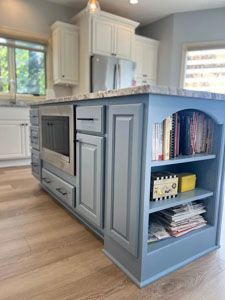 A blue kitchen island with a built-in microwave, cabinets, and open shelving filled with books on a light wood floor.