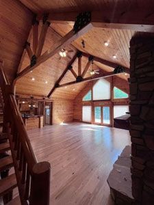 Spacious cabin interior with vaulted wooden ceilings, exposed beams, a stone fireplace, and light wood flooring.