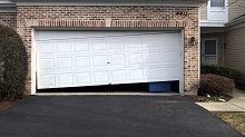 A white garage door is open and broken in front of a brick house.