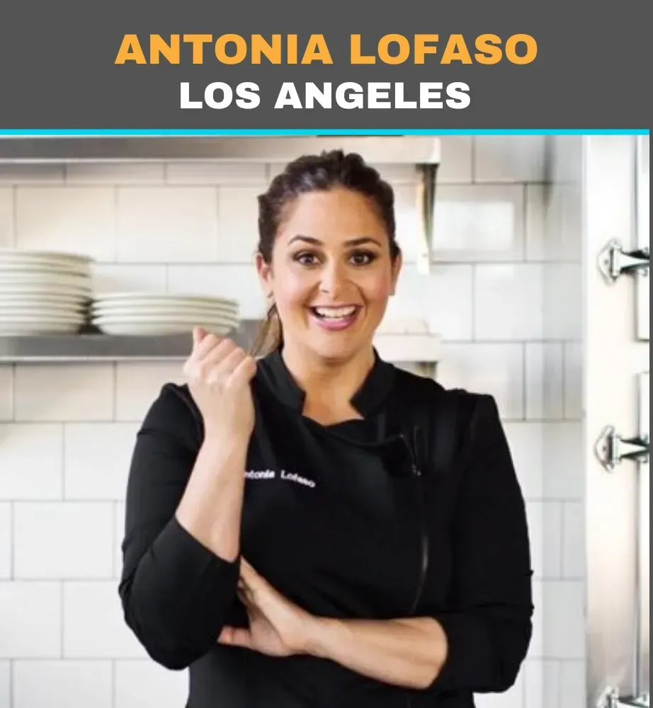 Antonia Lofaso smiling in a black chef coat in a bright kitchen, holding a spoon.