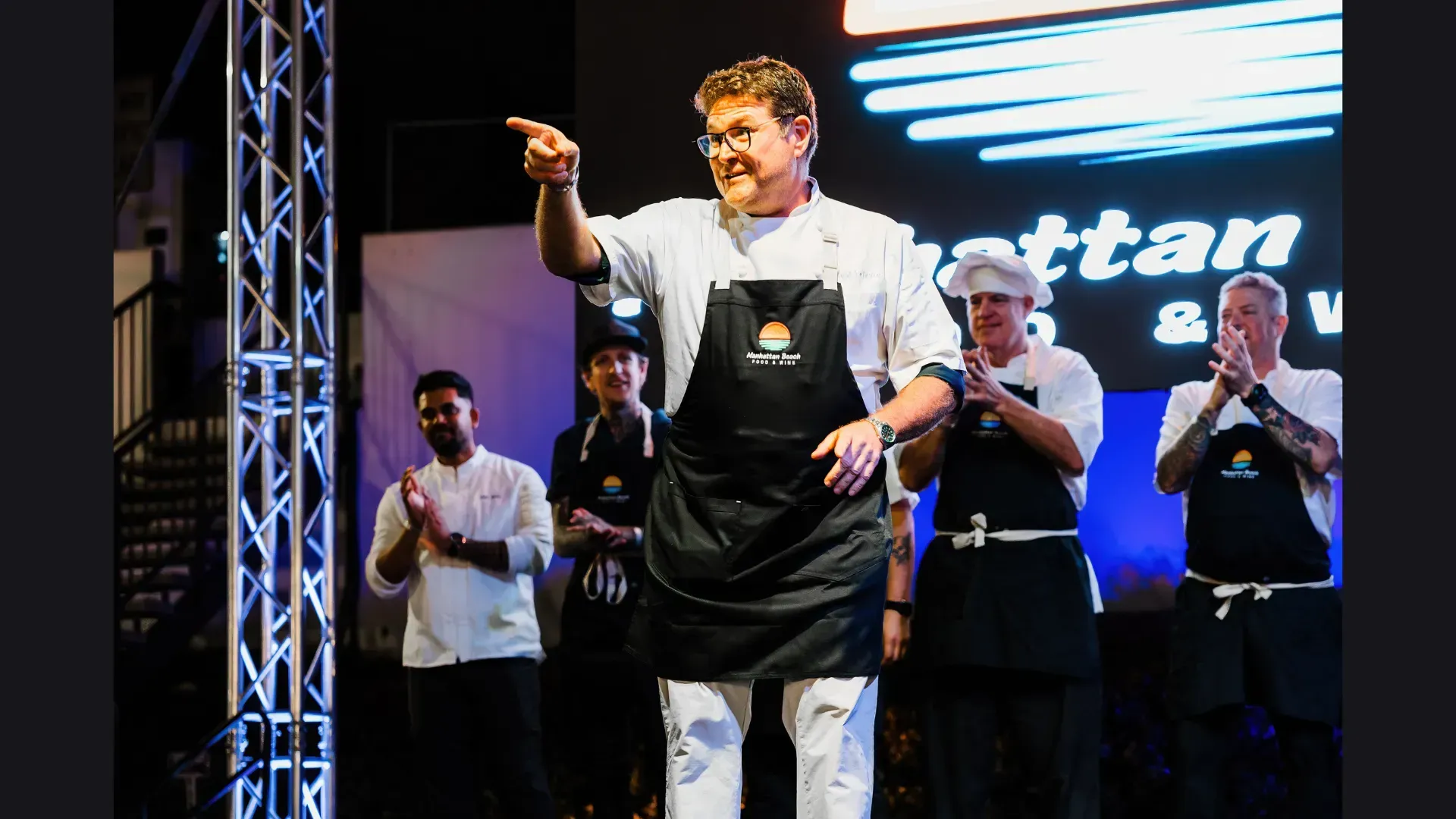 Man in black apron on stage points toward the audience, with people clapping behind him.