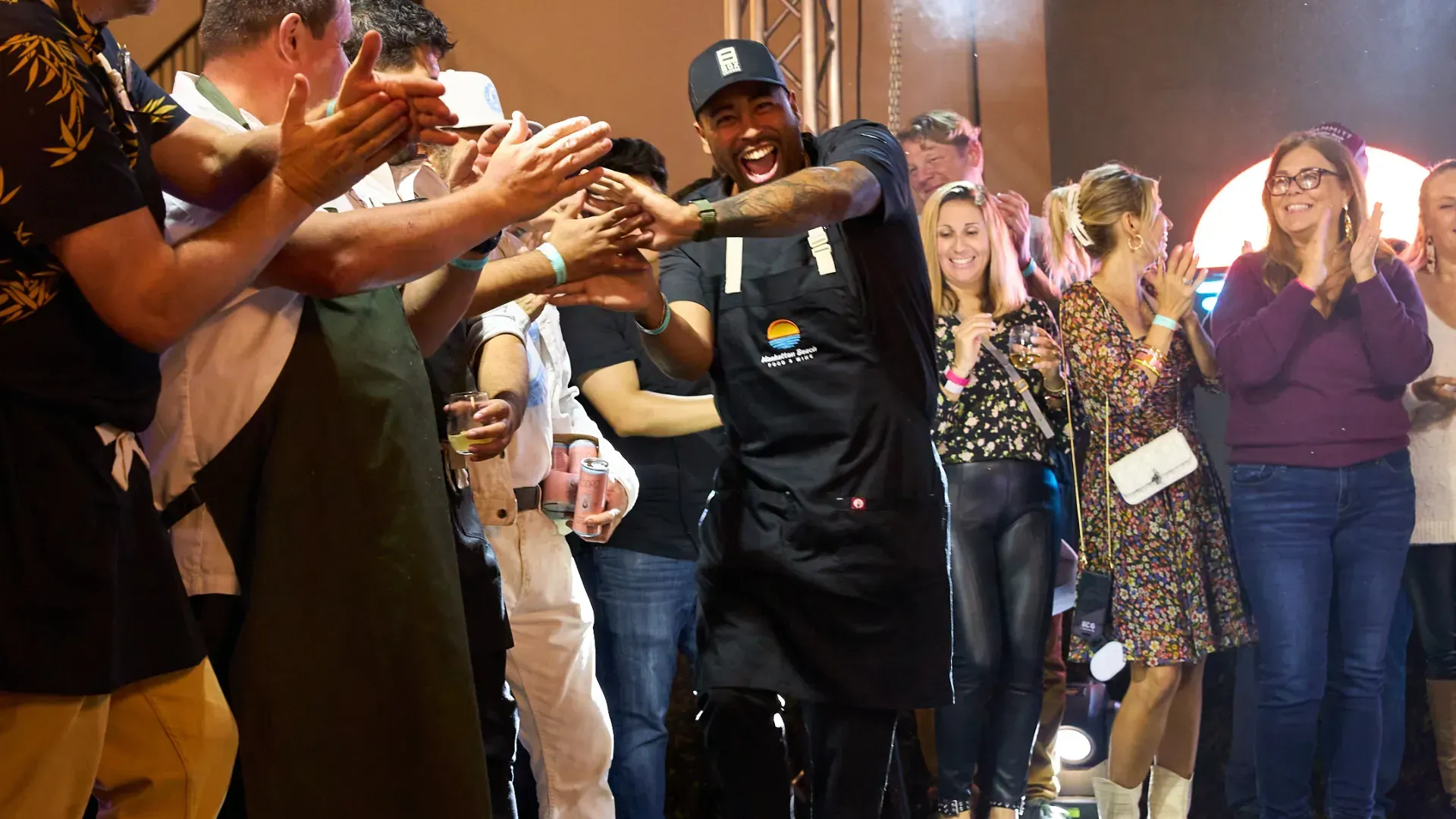 A crowd claps and reaches out as a man in black dances in the center of a lively indoor event.