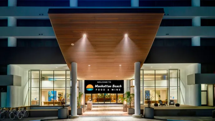 Modern hotel entrance at dusk with glass doors, warm wood canopy, and lit lobby sign reading “Pebble Beach Four Seasons.”