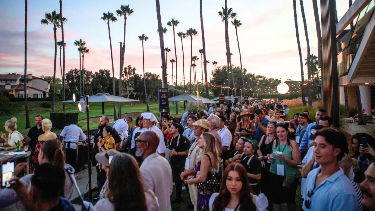Crowd gathered outdoors at sunset near palm trees and a building entrance, chatting and socializing