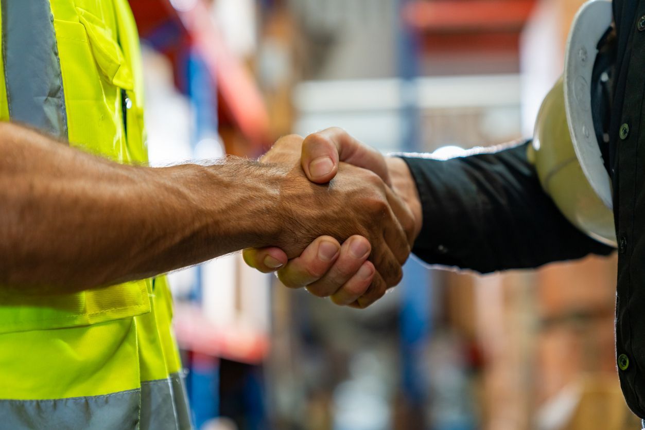 Two people shaking hands in a warehouse; one wearing a safety vest, the other holding a hard hat.