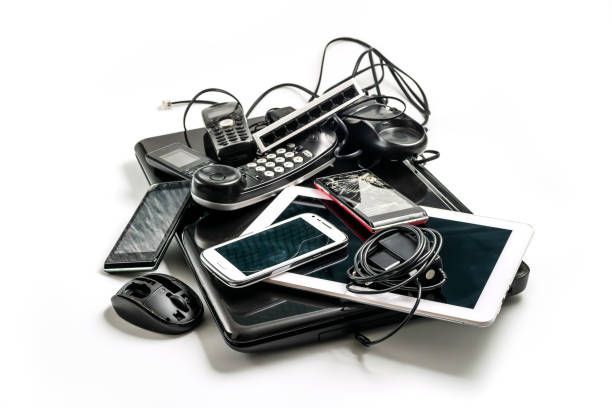 Pile of discarded electronics: phones, tablet, laptop, mouse, tangled cords, against a white background.
