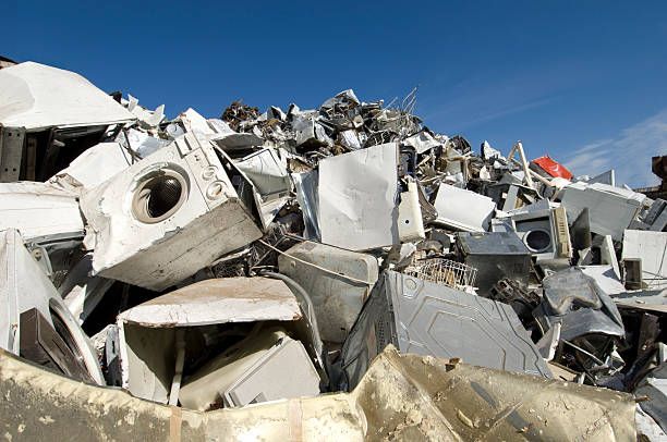 Pile of discarded white appliances against a blue sky, including washing machines, and dryers at a recycling facility.