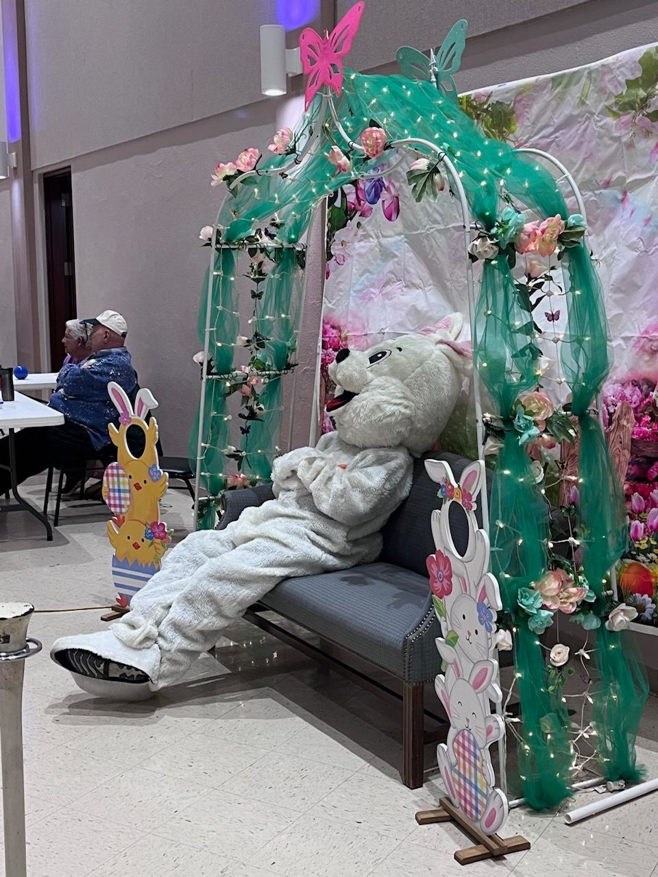 A teddy bear in a bunny costume is laying on a bench under a green arch.