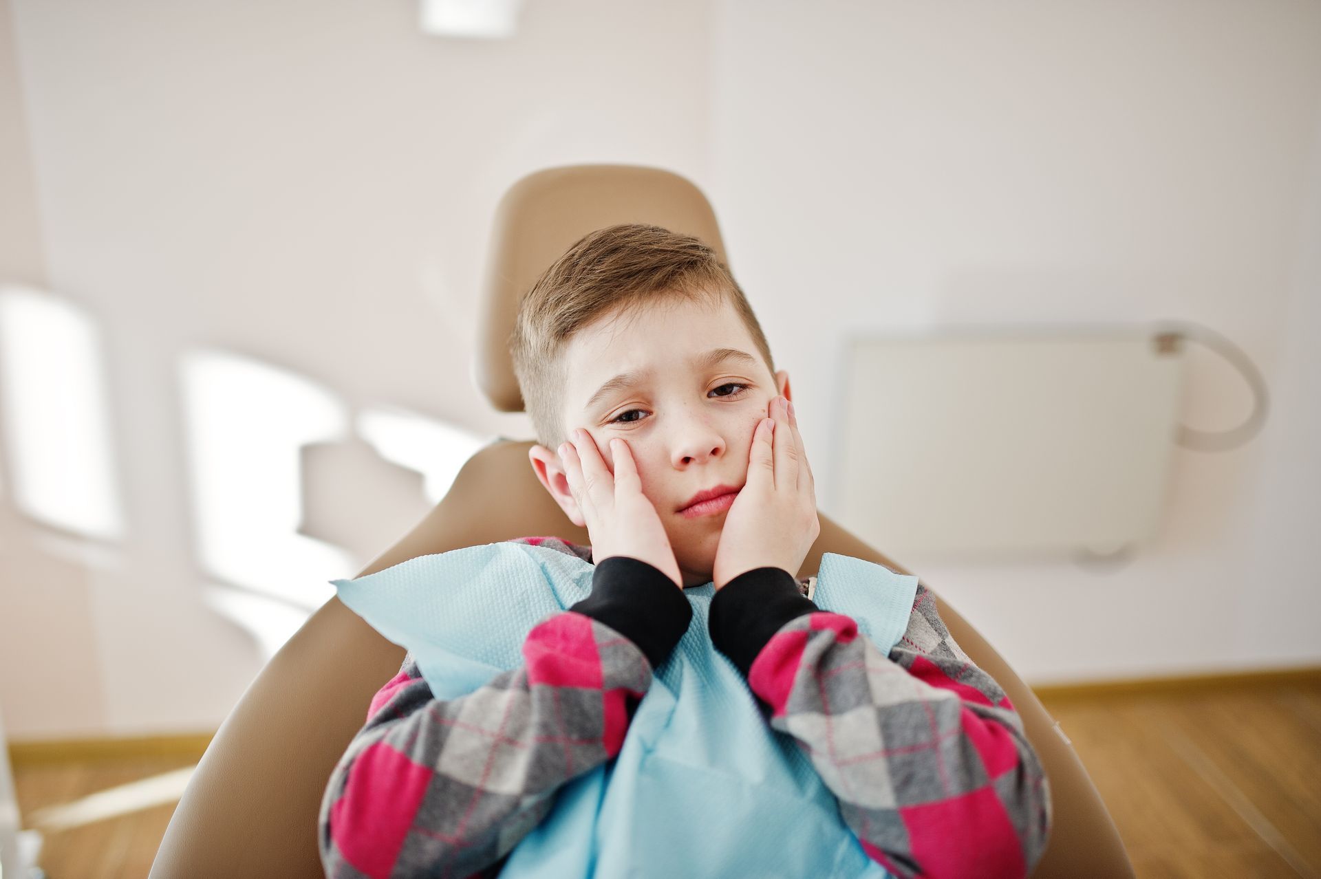 A young boy is sitting in a dental chair with his hands on his face.