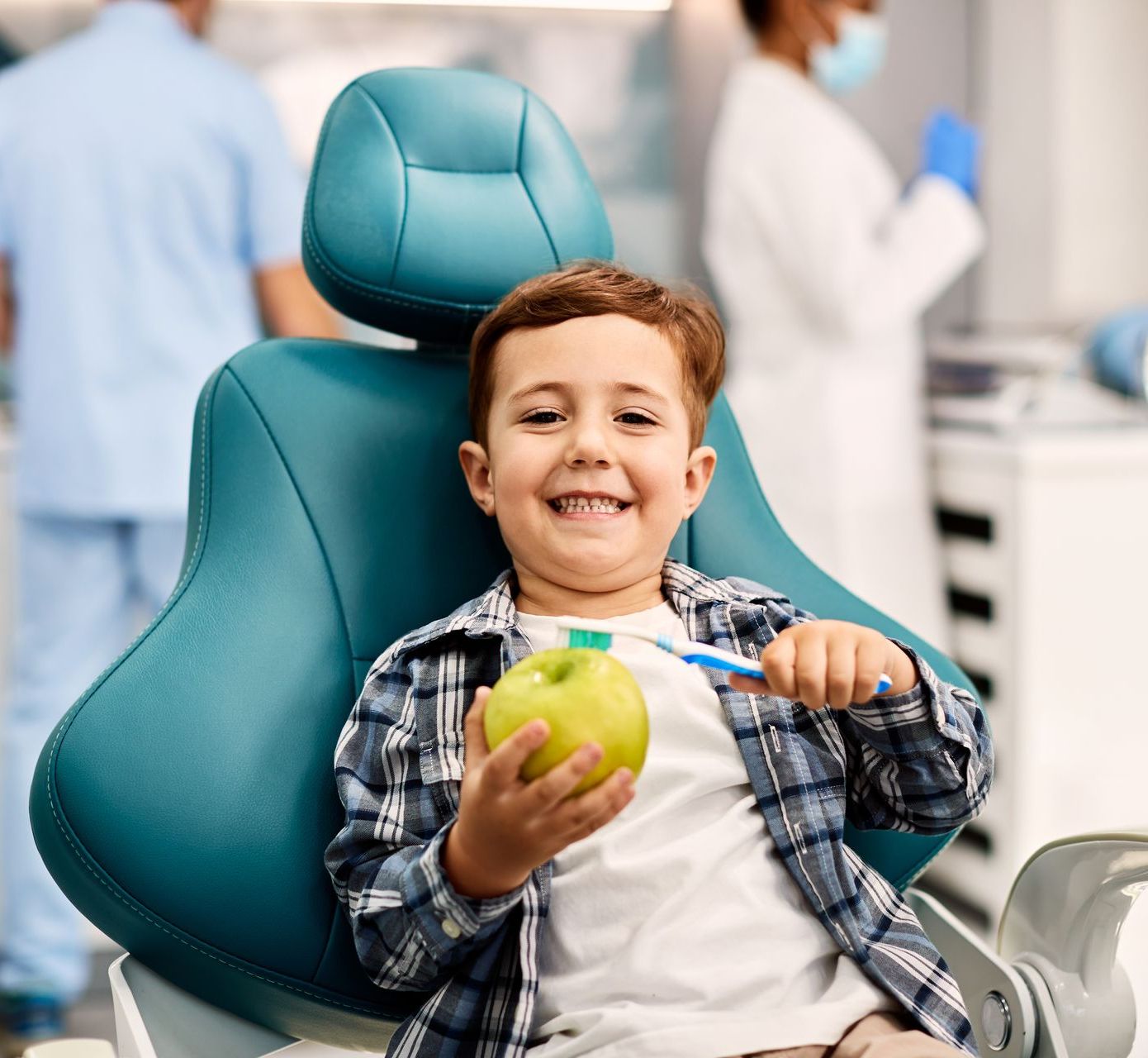 A young boy is sitting in a dental chair holding an apple and a toothbrush.