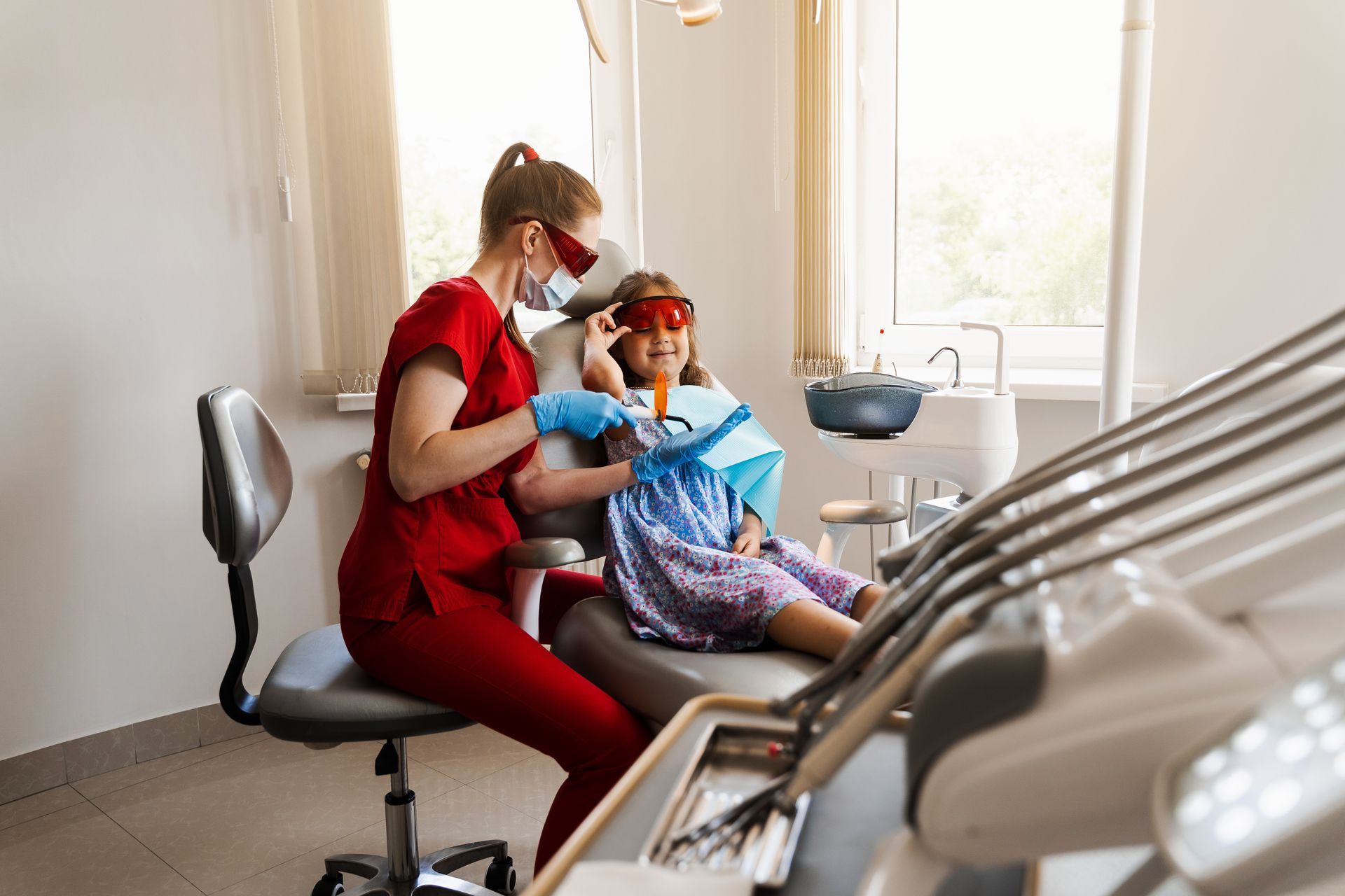 Dentist in red scrubs examining a child in a dental chair; bright light in the window.