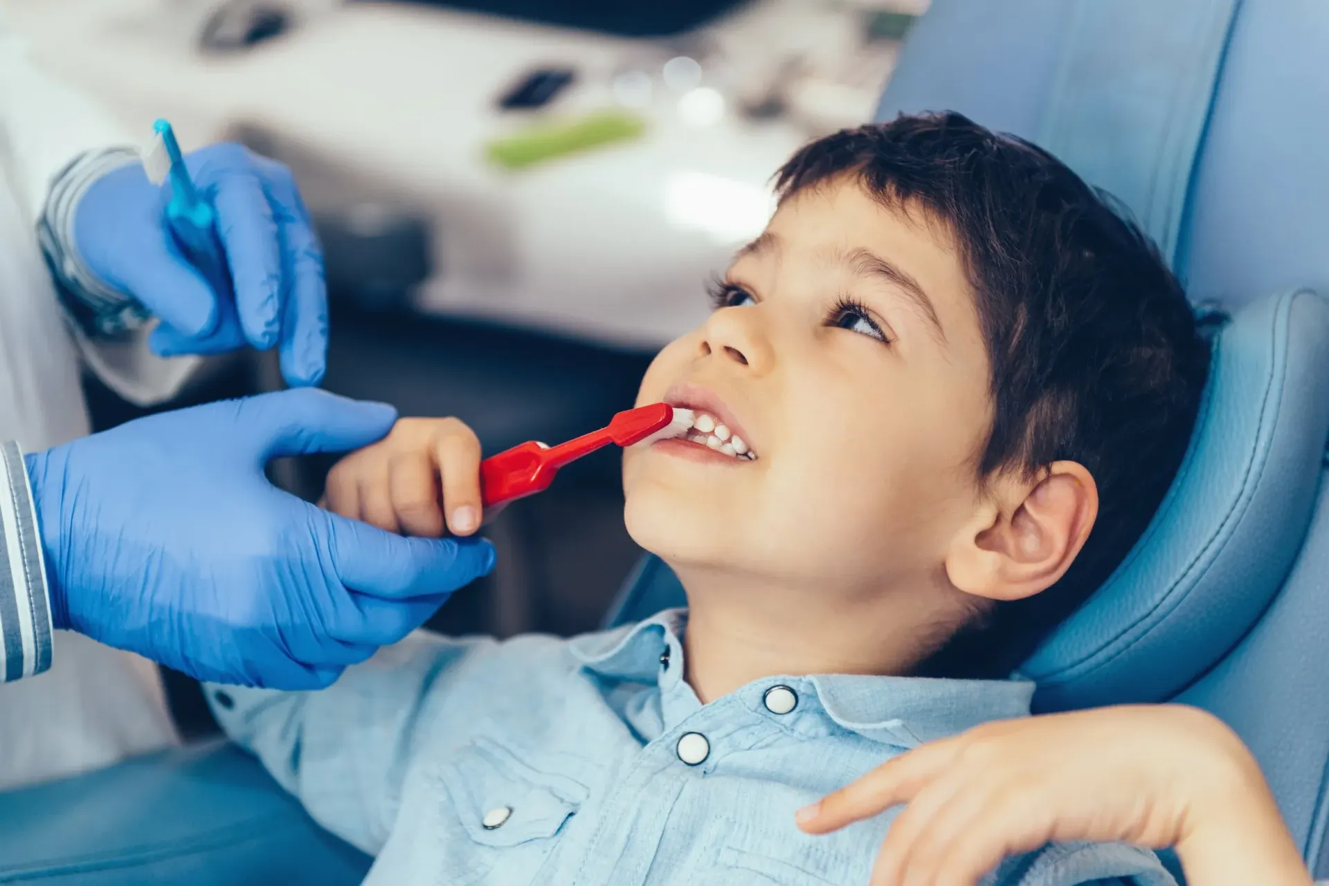 A dentist in blue gloves helps a young boy brush his teeth with a red toothbrush in a dental chair.