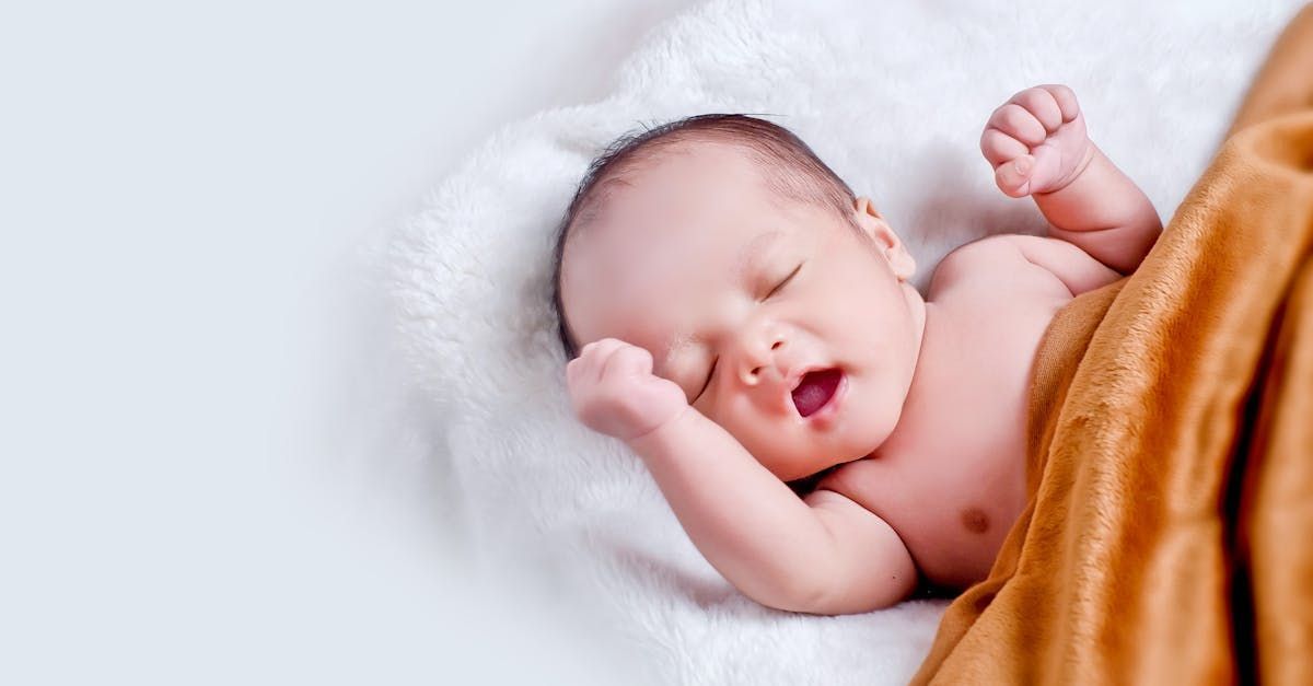 A newborn baby is sleeping on a white blanket with its mouth open.