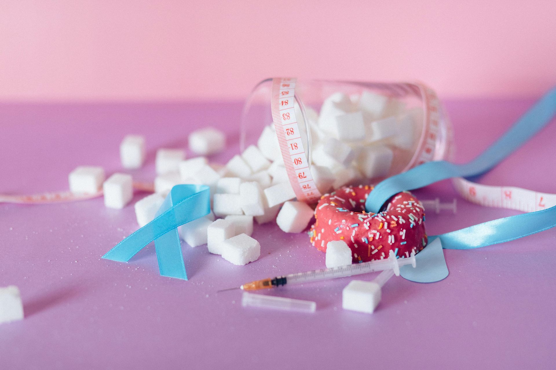 A donut , measuring tape , sugar cubes and a syringe on a pink table.