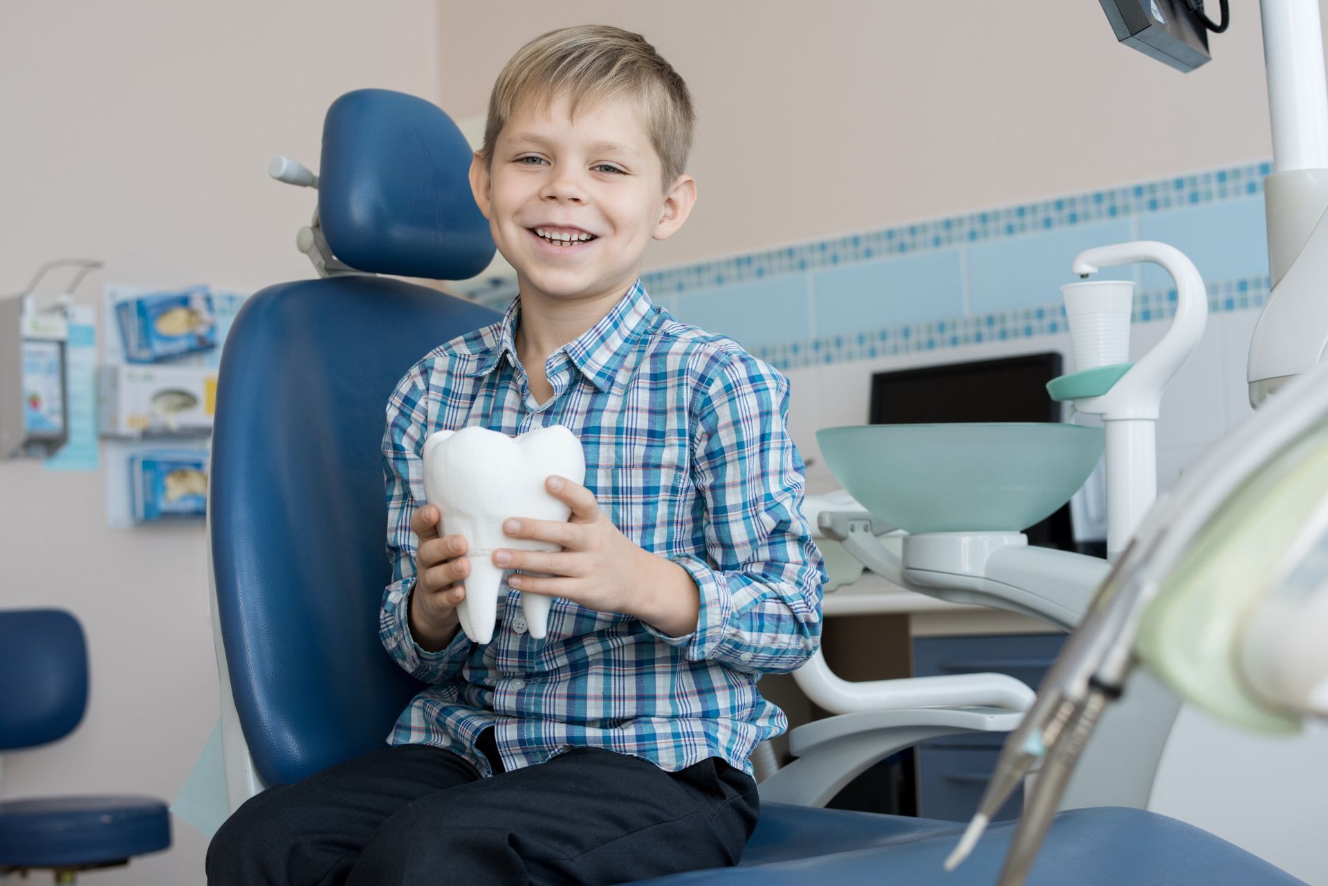 A young boy is sitting in a dental chair holding a model of a tooth.