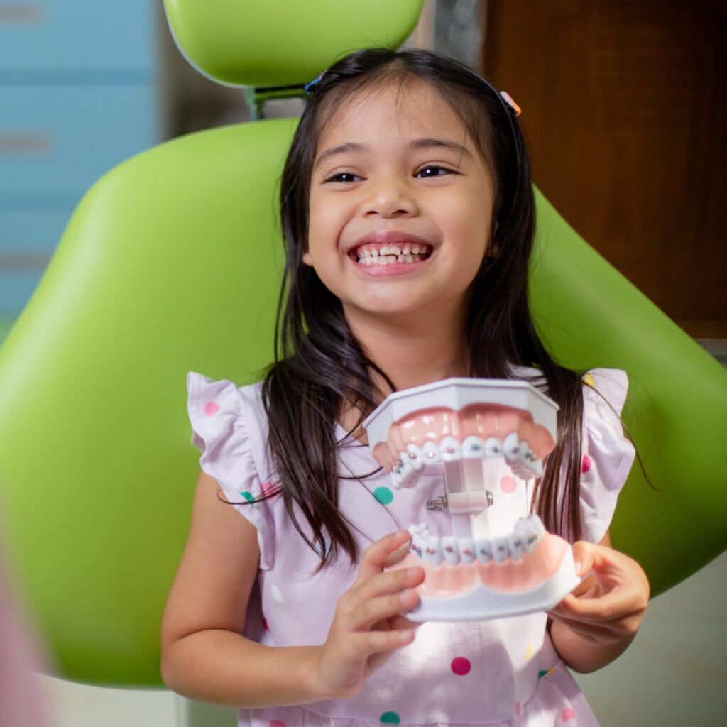 A little girl is sitting in a dental chair holding a model of her teeth.