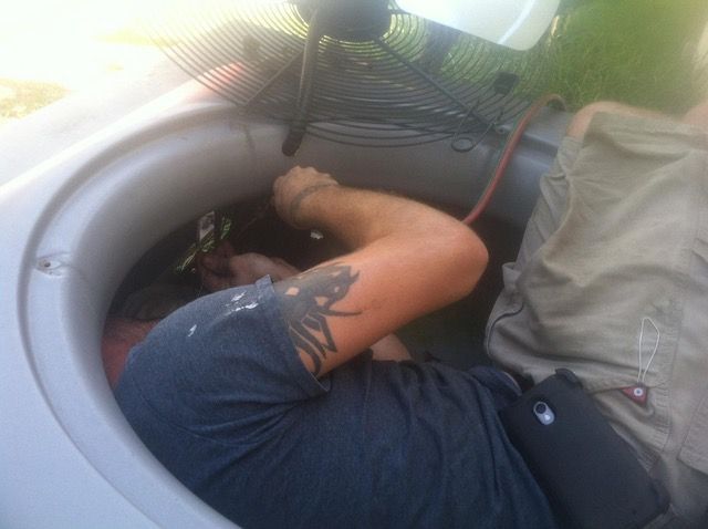 Man working on an air conditioning unit; lying inside, using tools, with a fan above him.