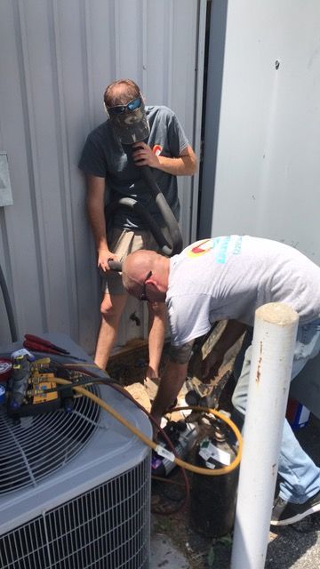 Two men work on an AC unit outdoors; one wears a face covering.