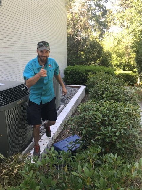 HVAC technician giving a thumbs up next to an air conditioning unit and bushes.