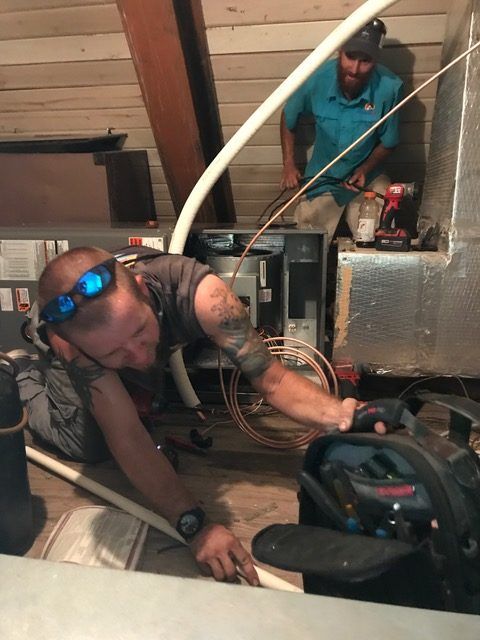 Two technicians working on HVAC equipment in an attic. One holds a tool, the other works near the unit.