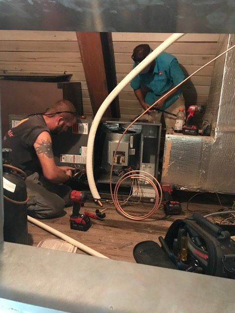 Two people installing HVAC equipment in an attic. One kneels, the other works standing. Tools and copper tubing are visible.