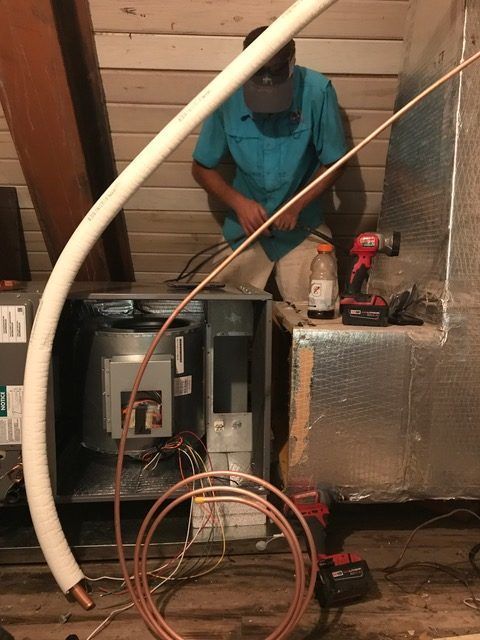A person working on HVAC system components in an attic, with tools and copper tubing visible.