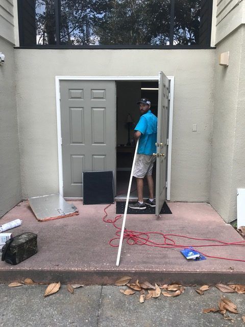 Man in doorway, holding a white pole. Outdoor setting with open door, building, electrical cords, and debris.