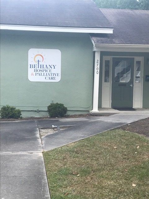 Exterior of Bethany Hospice & Palliative Care building; green facade, white sign, sidewalk, shrubs, and door.
