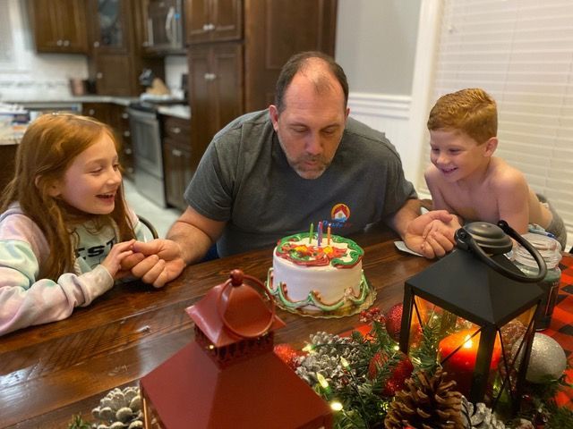 Man blowing out candles on a cake with two children at a wooden table, festive decorations.