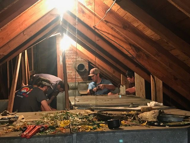 Men working in a dimly lit attic. They are repairing something near a brick chimney.