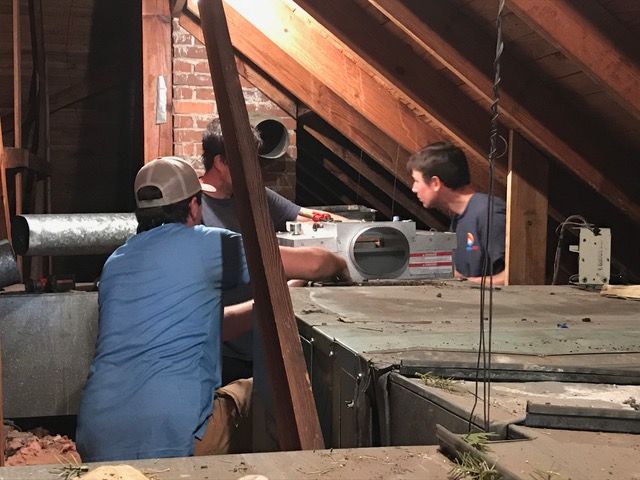 Three people working on HVAC equipment in an attic.