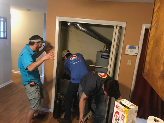 Three HVAC technicians working on a unit in a closet while another gestures nearby.