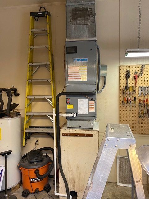 A furnace unit with a yellow ladder in a garage setting, near a water heater and tools on a pegboard.