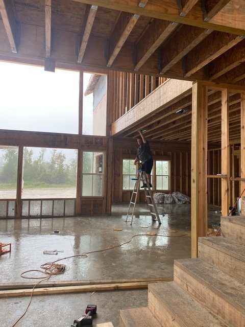 Construction worker on a ladder in a house frame. Interior with concrete floor and large windows overlooking outdoors.