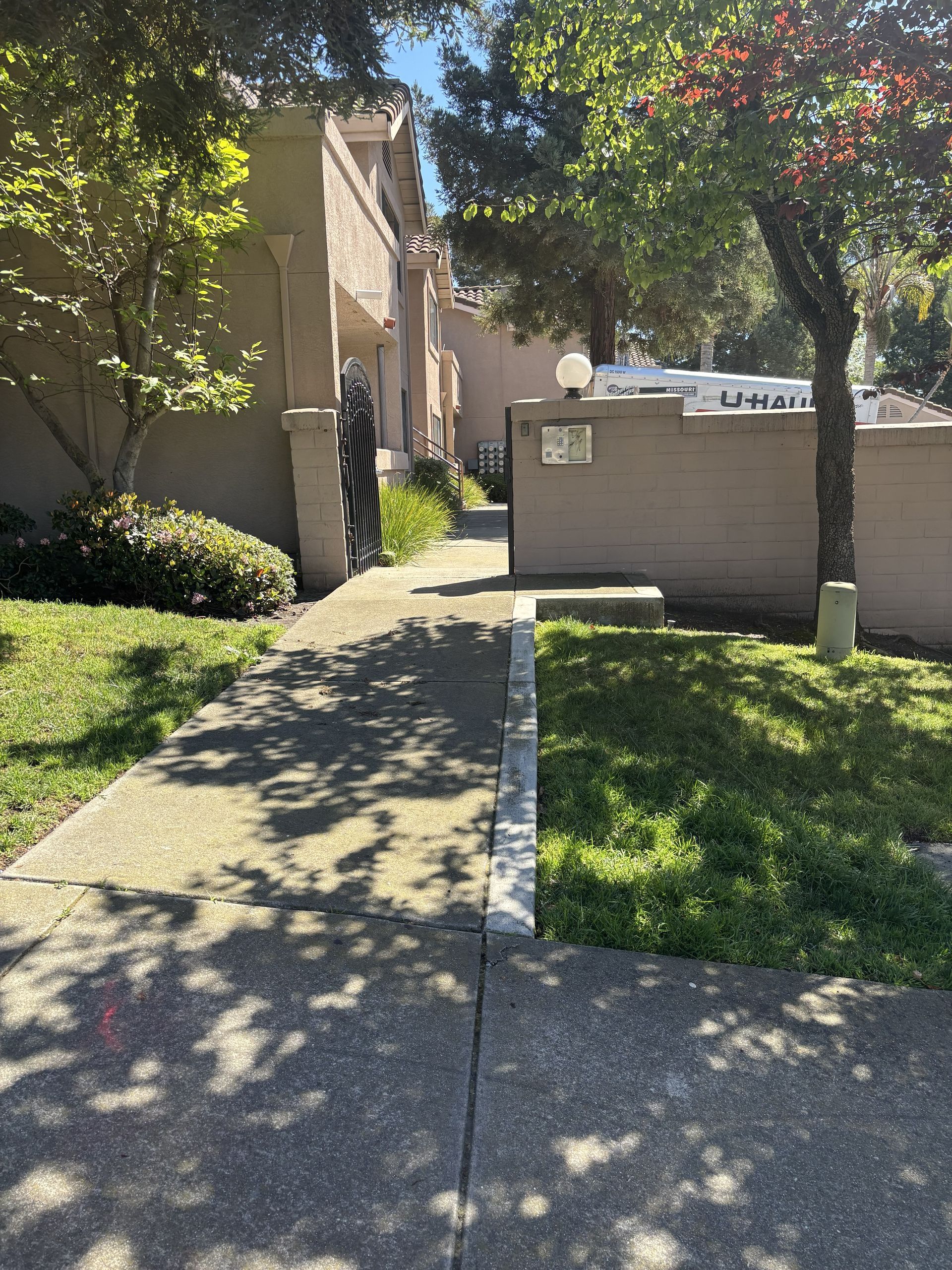 A concrete path leads between a beige building and a garden wall, dappled with tree shadows on a sunny day.