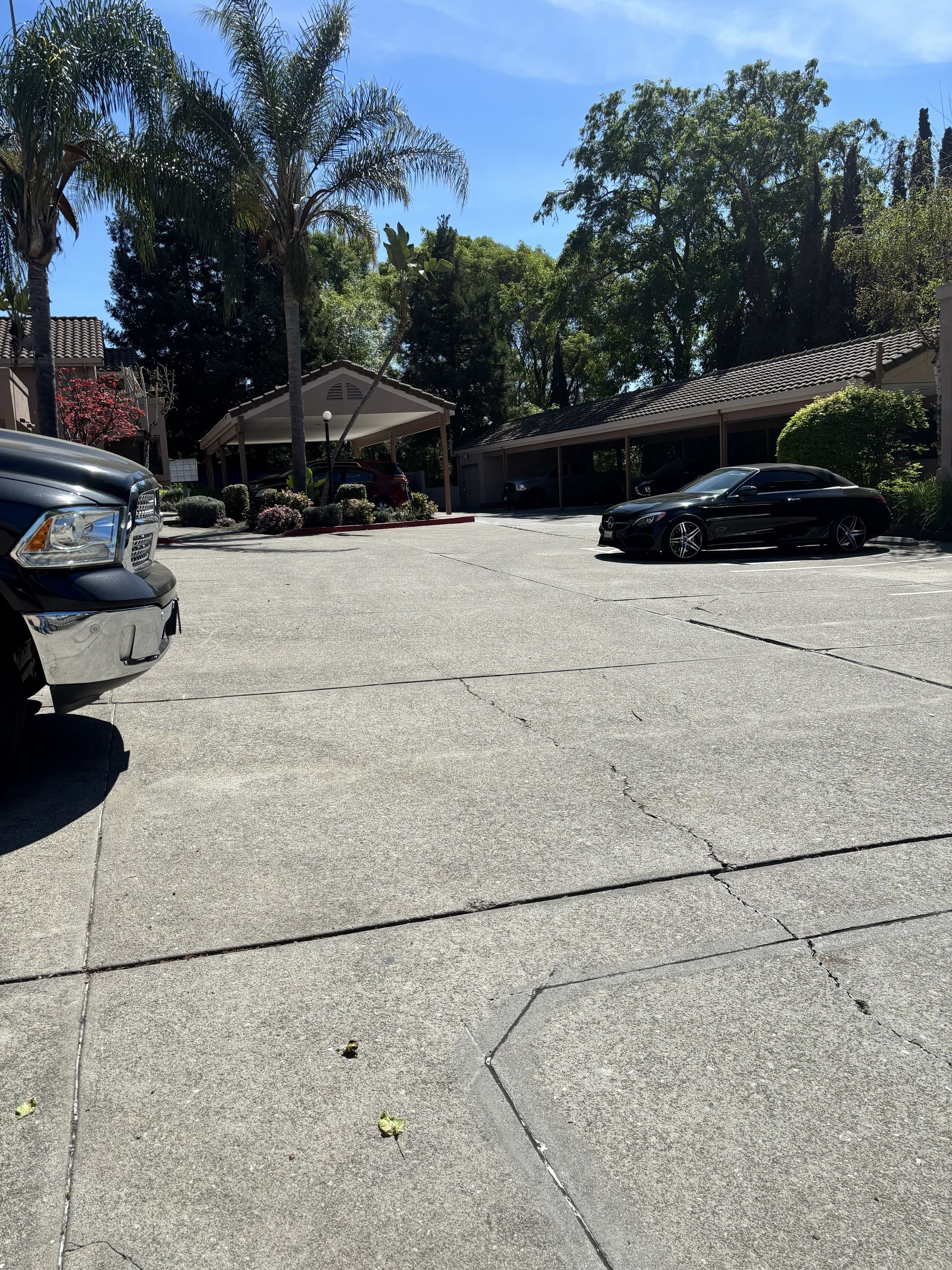 A sunlit parking lot with a pickup truck in the foreground and a black sedan parked under a covered structure.